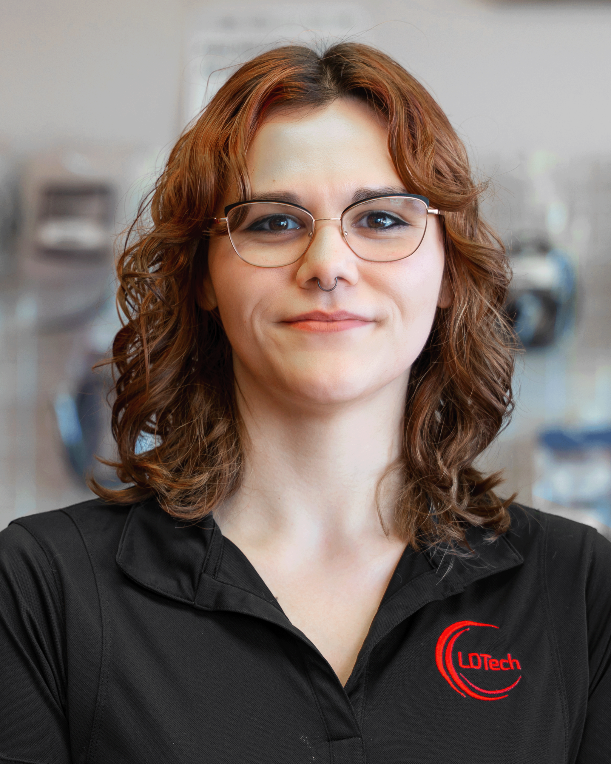 Close-up portrait of a smiling woman with auburn wavy hair, round glasses, and a nose ring in a black polo with a red logo.