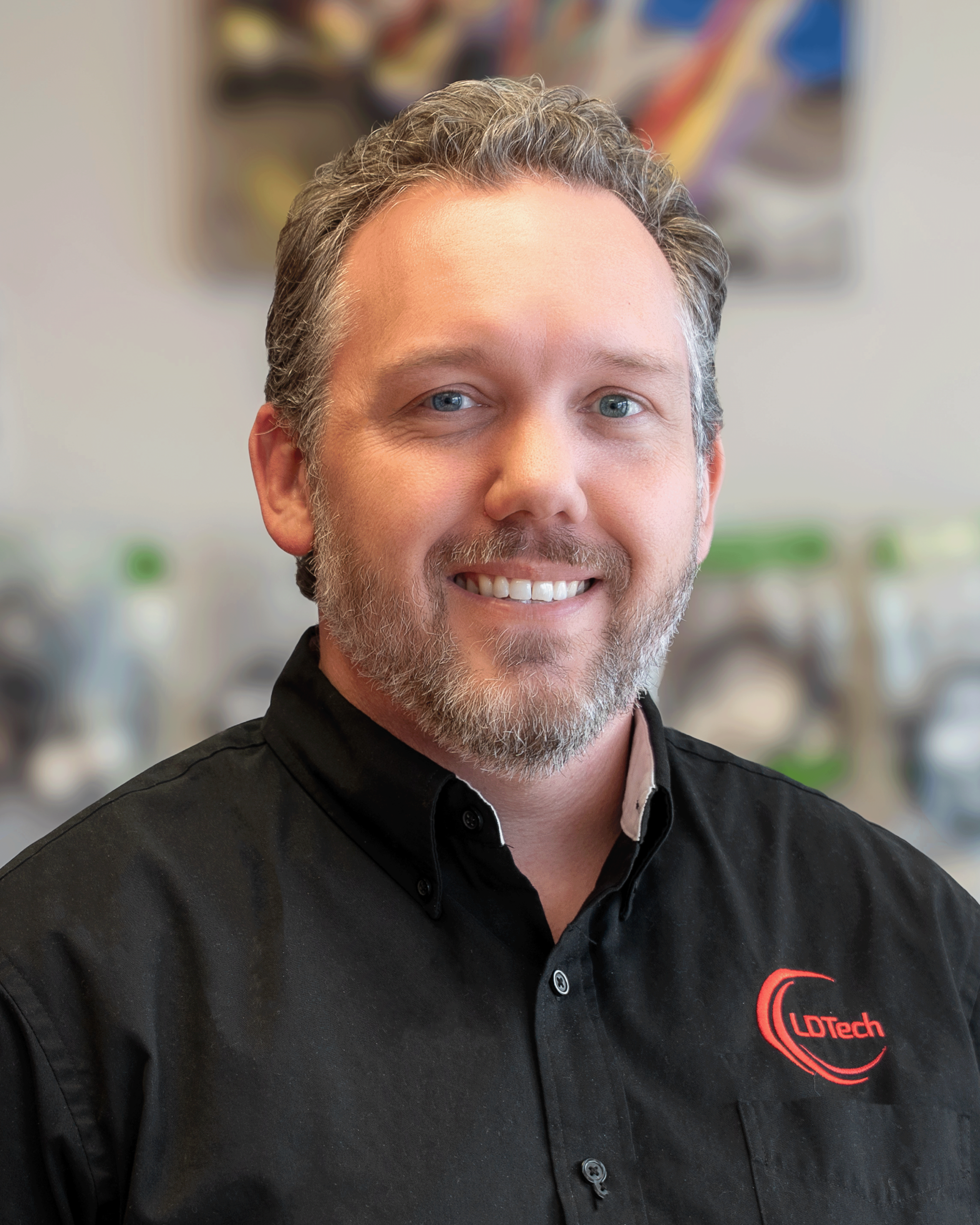 Close-up portrait of a smiling man with gray hair and beard wearing a black shirt with an LDTech logo on the chest
