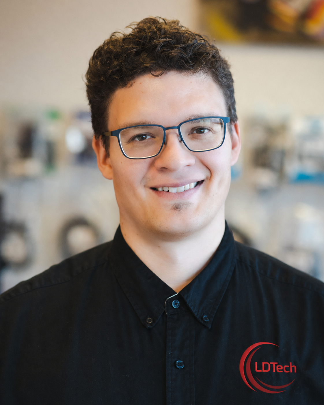 Smiling man wearing blue glasses and a black LDTech polo shirt, posed for a portrait in a tech office.