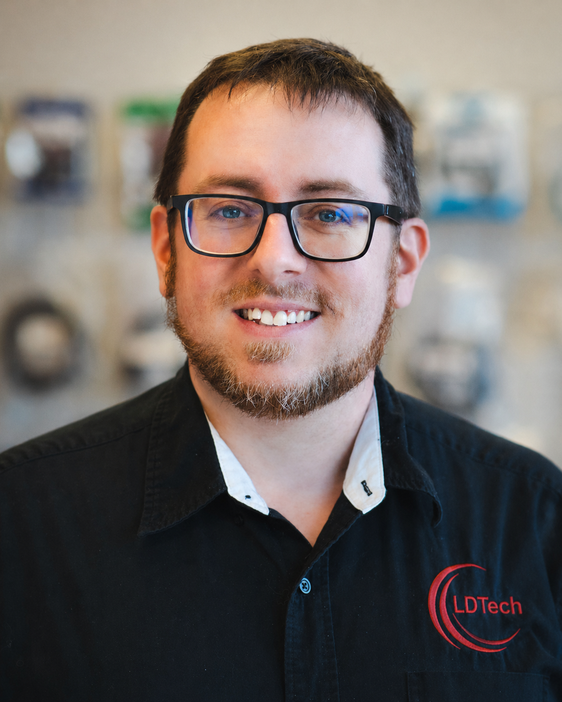 Smiling man with short dark hair and glasses, wearing a black LDTech polo, in a blurred indoor background.