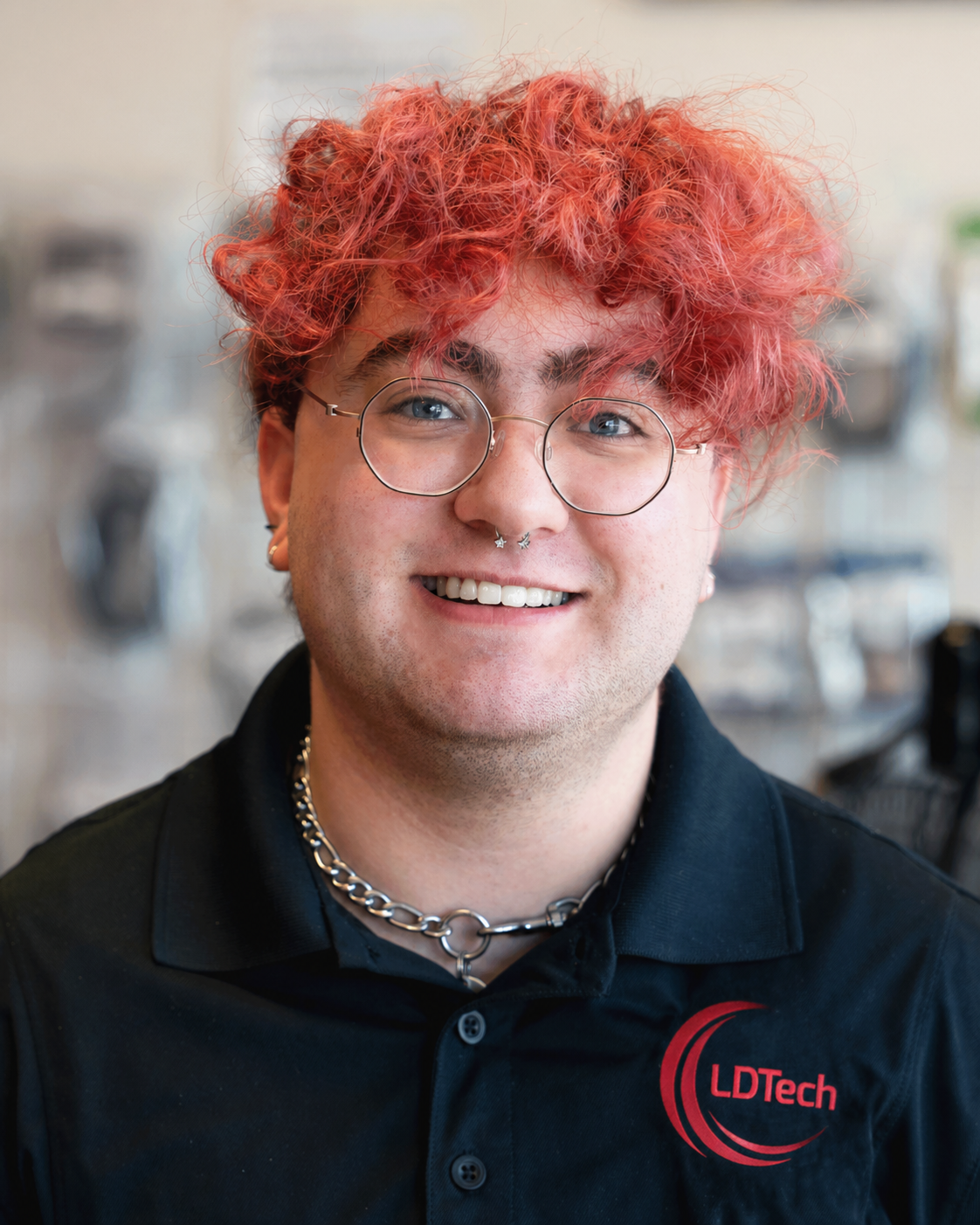Person with bright red curly hair and round glasses smiling at the camera in a black LDTech polo shirt.