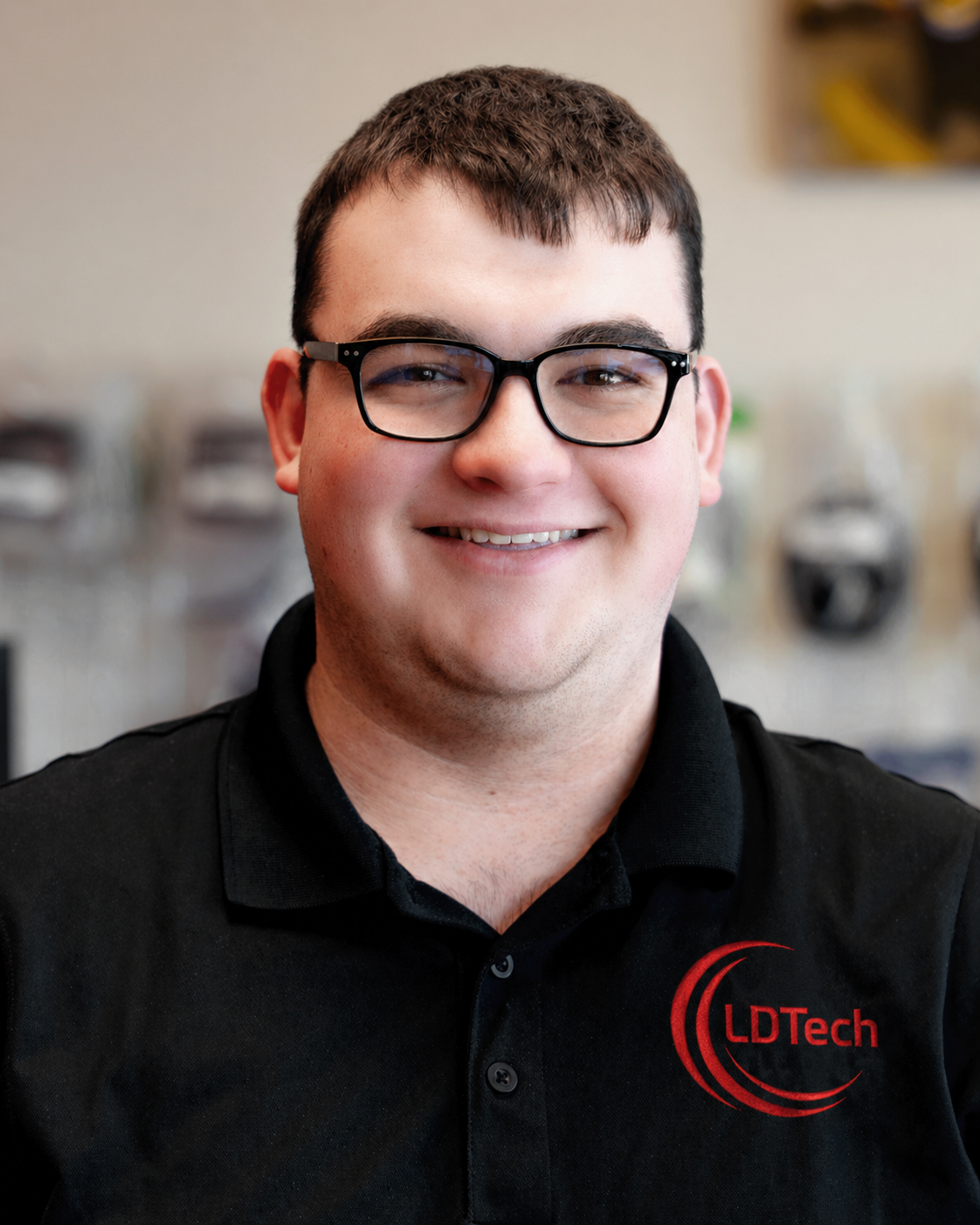 Smiling man with short dark hair and black rectangular glasses, wearing a black polo shirt with an LDTech logo in red on the chest, indoors.